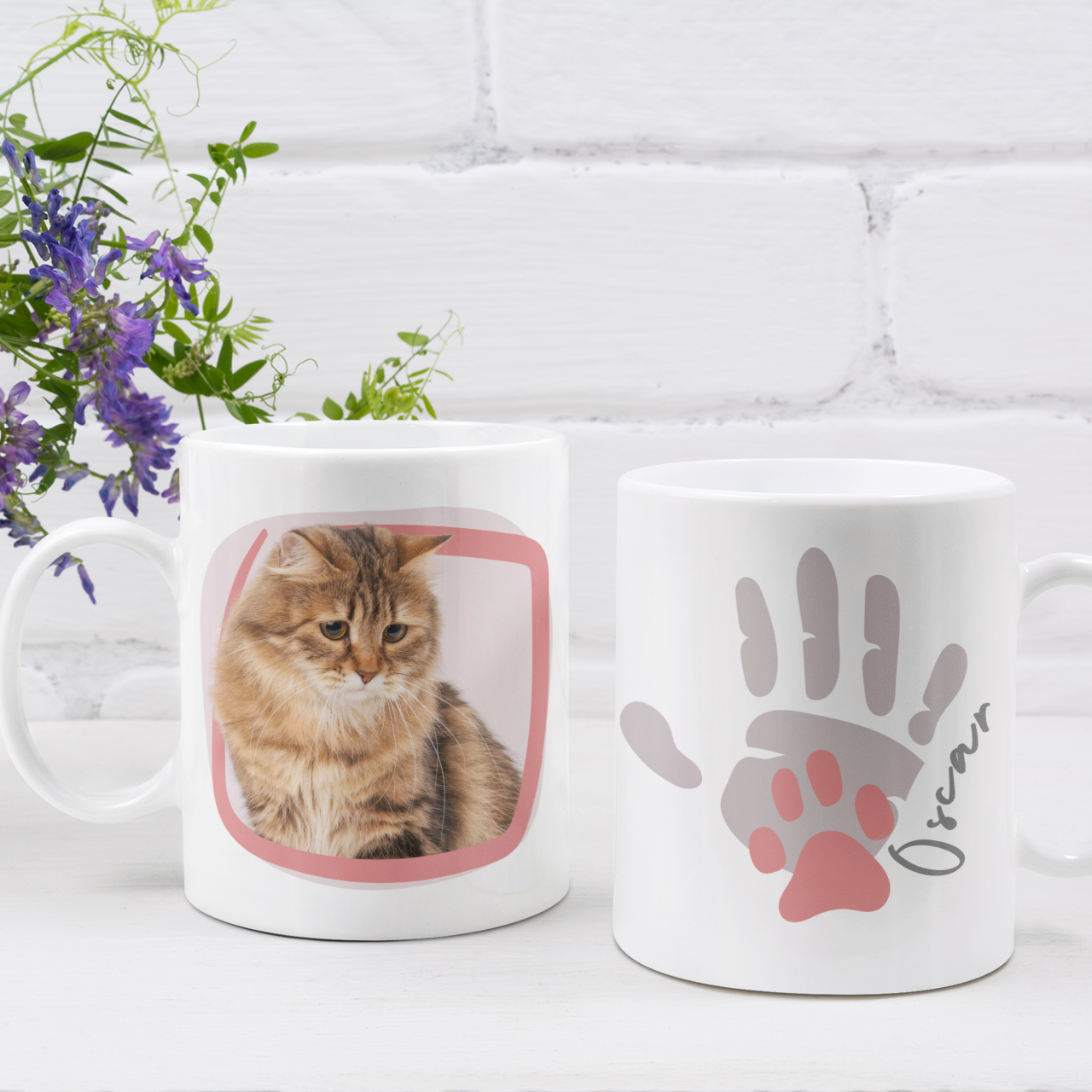 Two mugs with a cat design and paw print on a white surface with a plant in the background.